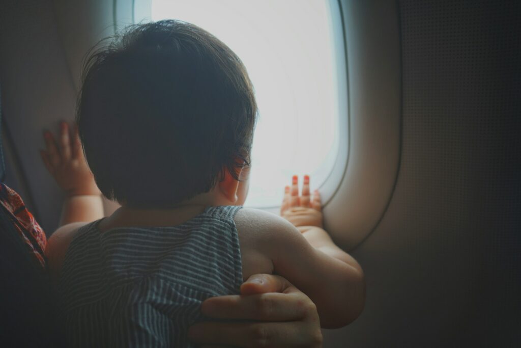 The back of a toddler's head as it peers out an airplane window with a parent's arm wrapped around its middle, representing hague convention and child abductions.