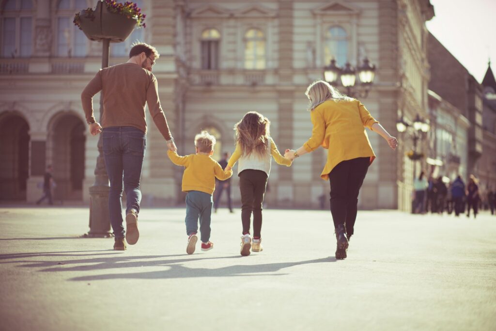 A family with two young children walks along a street path with an old building in front of them, representing a new relationship after divorce.